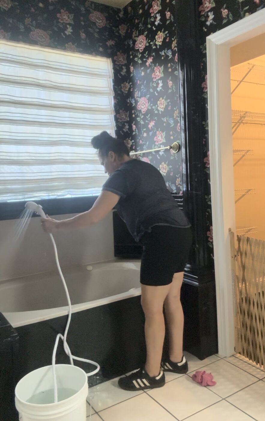 Angela cleaning a bathtub by hand with a spray hose, mid-job.